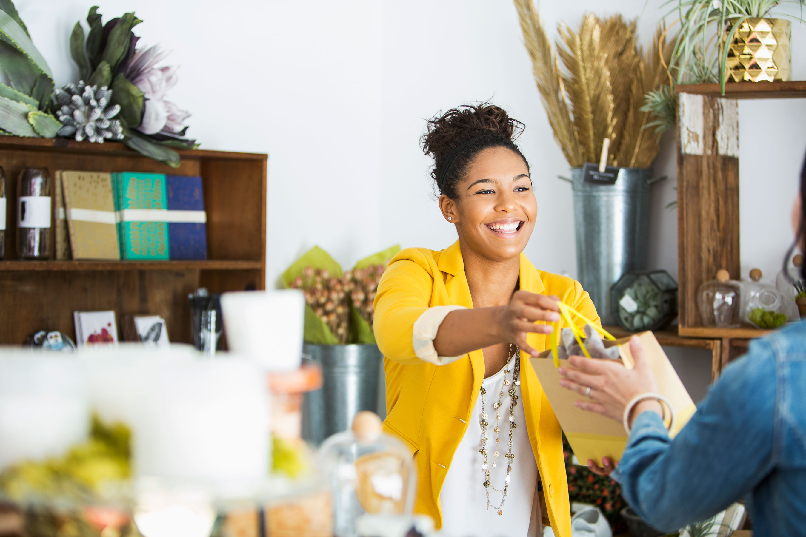 Woman Wearing Yellow, Smiling and Handing a Customer Their Bag in Boutique Store